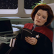 Captain Janeway sits at her desk in her ready room, coffee mug in one hand, PADD in the other.